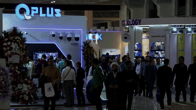Diversas pessoas caminham e conversam em corredor de feira de tecnologia. Estandes iluminados exibem nomes como QPlus e outros, com arranjos florais decorativos ao lado de alguns estandes.