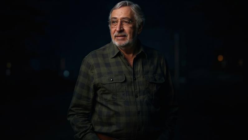 Homem idoso com barba e óculos usa camisa xadrez verde escura e calça jeans, com as mãos nos bolsos, em área externa urbana com prédios desfocados ao fundo.