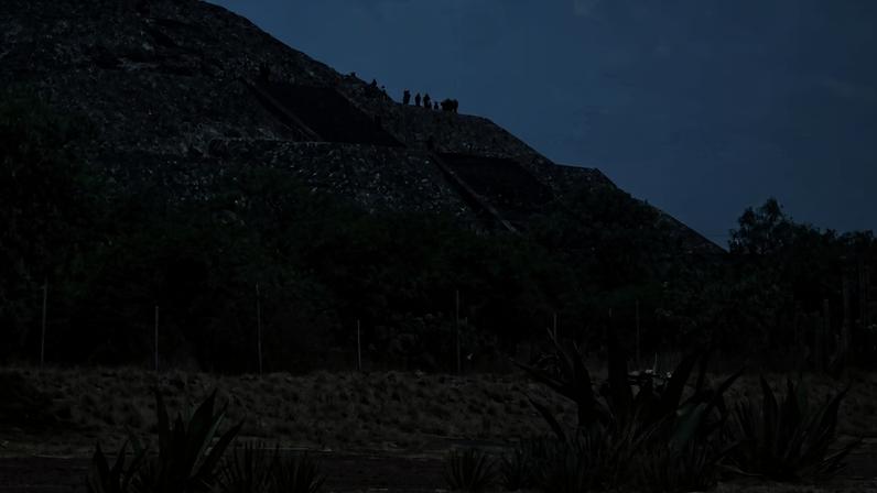 Pirâmide de pedra com escadaria central e pessoas no topo. Vegetação rasteira e arbustos cercam a base da estrutura sob céu claro.