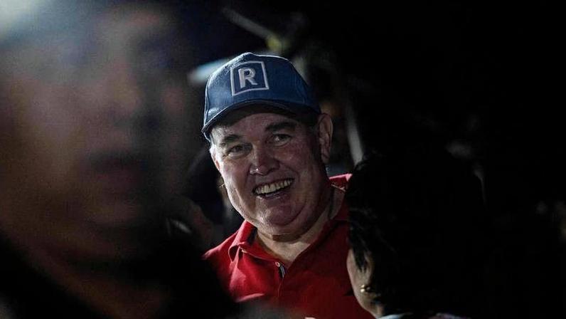 Homem de camiseta vermelha e boné azul com a letra 'R' branca sorri em ambiente escuro, cercado por outras pessoas parcialmente visíveis.