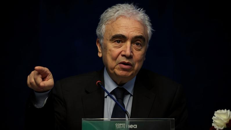 Homem de cabelos grisalhos e expressão séria fala em conferência, usando terno preto e gravata azul. Ele gesticula com a mão direita enquanto está atrás de um púlpito com placa que indica o nome 'Mr. Fatih Birol'. Fundo azul claro desfocado.