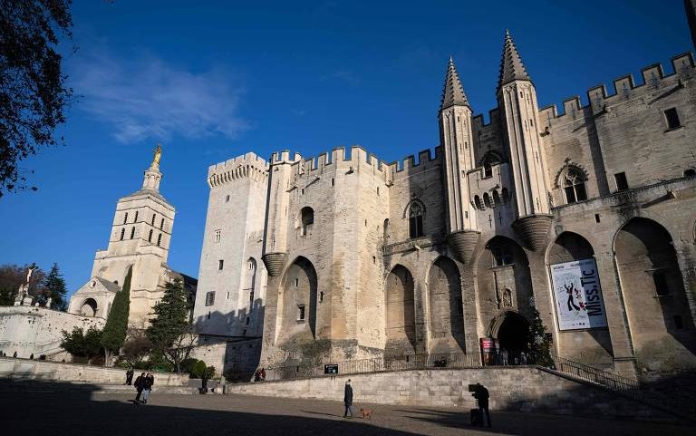 Palácio dos Papas em Avignon com arquitetura medieval em pedra clara, torres e muralhas sob céu azul. Pessoas caminham na praça em frente.