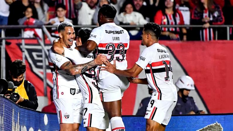 Quatro jogadores do São Paulo em uniforme branco com detalhes vermelhos e pretos comemoram gol juntos na beira do campo. Um jogador é erguido pelos companheiros enquanto a torcida ao fundo vibra nas arquibancadas.