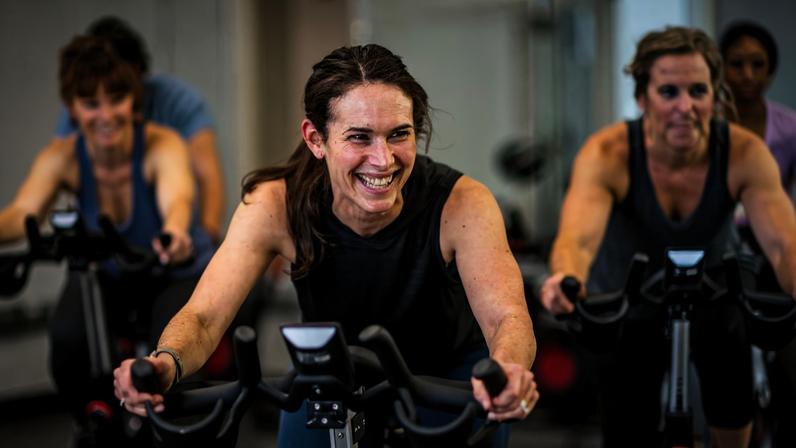 Cinco pessoas pedalando em bicicletas ergométricas em ambiente interno de academia. Mulher no centro sorri enquanto pedala, outras ao redor focadas no exercício. Ambiente iluminado com paredes claras e equipamentos modernos.