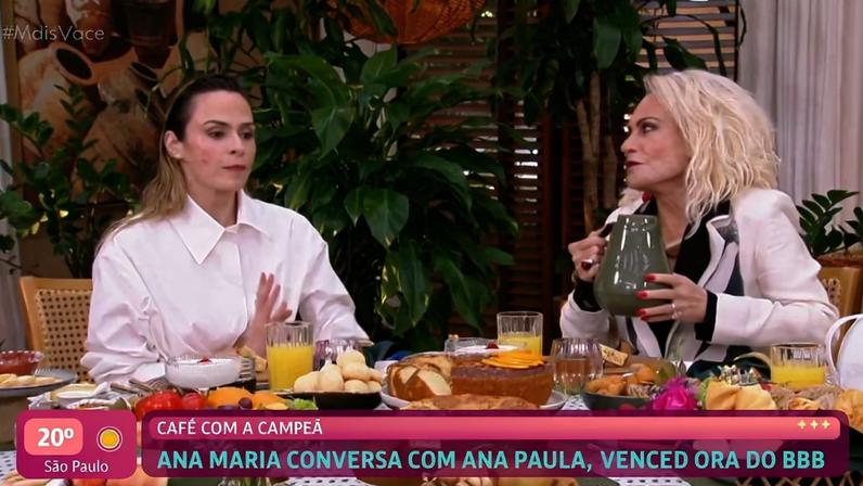 Ana Maria Braga e Ana Paula, vencedora do BBB, sentadas à mesa com variedade de pães, frutas, sucos e café, em ambiente interno com plantas ao fundo.