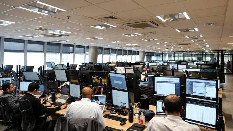 Sala ampla de escritório com fileiras de mesas equipadas com múltiplos monitores. Pessoas sentadas trabalham em computadores, algumas usando fones de ouvido. Iluminação fluorescente no teto e janelas grandes ao fundo fornecem luz natural. Ambiente moderno e organizado, com cadeiras ergonômicas e equipamentos tecnológicos.