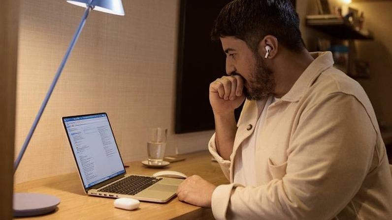 Homem sentado à mesa em ambiente interno, usando fones de ouvido sem fio e olhando para a tela de um laptop aberto. Ao lado do laptop há um copo de água e uma caixa de carregamento para os fones. Uma luminária azul está posicionada à esquerda, iluminando a área de trabalho.