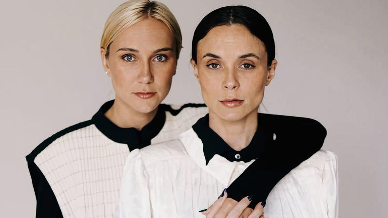 Duas mulheres estão próximas, uma atrás da outra, com a mulher de trás envolvendo o braço ao redor da frente da outra. Ambas usam roupas predominantemente brancas com detalhes pretos, em fundo neutro.