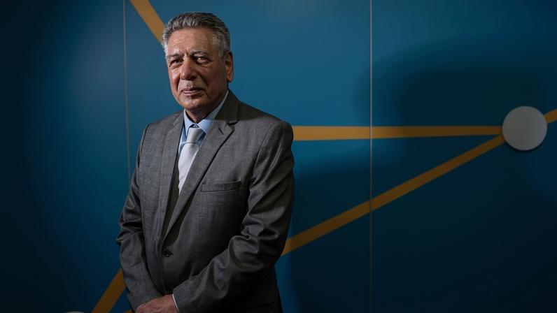 Homem de terno cinza e gravata azul claro está em pé diante de parede azul com linhas amarelas e círculos cinza, olhando para a câmera.