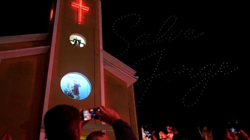 Fachada de igreja iluminada com cruz vermelha e projeção de cavaleiro medieval. No céu noturno, drones formam as palavras 'Salve Jorge'. Pessoas no primeiro plano registram a cena com celulares.