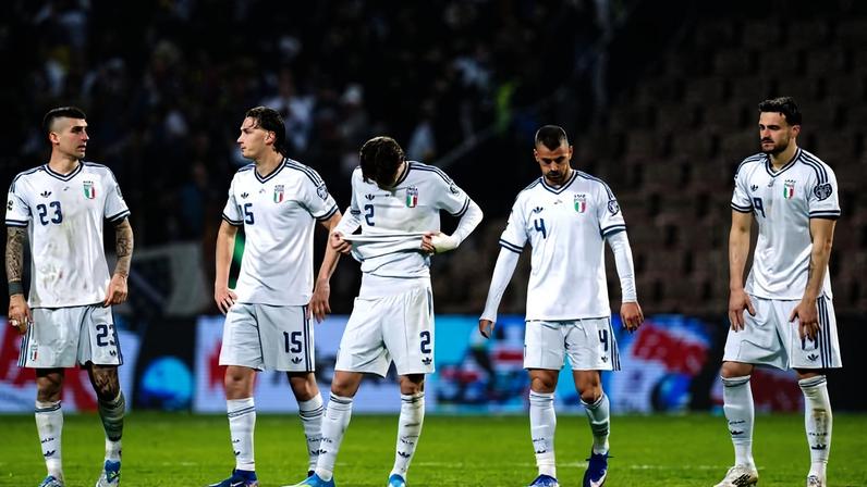 Cinco jogadores da seleção italiana de futebol em uniforme branco alinhados no campo, com expressão séria. Público desfocado ao fundo e arquibancada parcialmente vazia à direita.