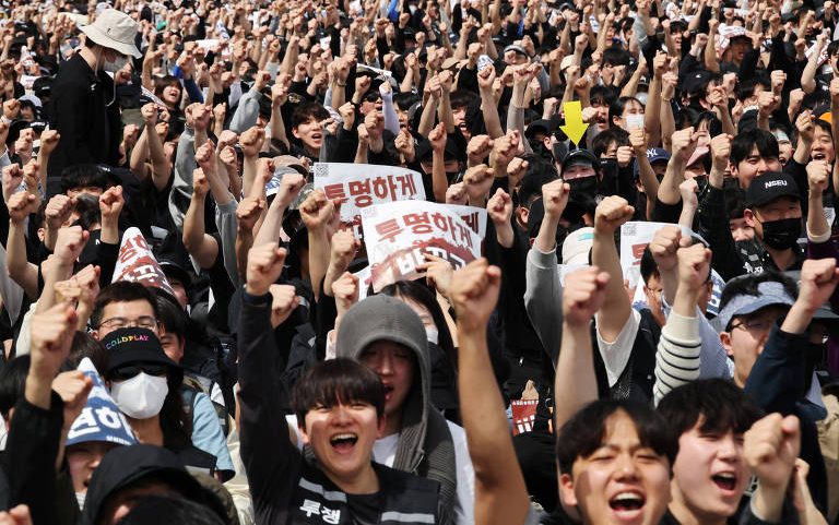 Centenas de pessoas reunidas em protesto, muitas com punhos erguidos. Algumas seguram cartazes com texto em coreano. A maioria usa roupas casuais e algumas usam máscaras faciais.