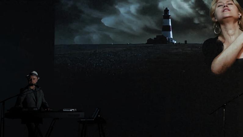Músico toca teclado à esquerda em ambiente escuro. Ao fundo, projeção mostra farol em paisagem noturna e rosto de mulher cantando com olhos fechados.