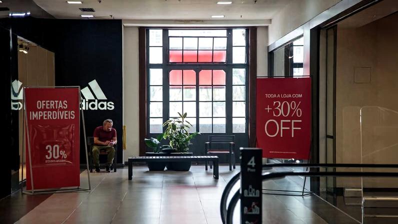 Interior de shopping com loja Adidas ao fundo. Dois cartazes vermelhos anunciam 30% de desconto em toda a loja. Homem sentado próximo à entrada, planta decorativa no centro.