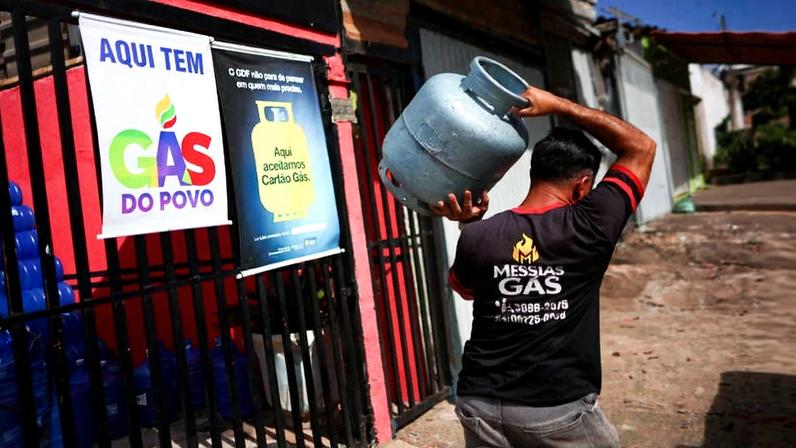 Homem de costas carregando um botijão de gás azul no ombro, caminhando em calçada ao lado de portão com dois cartazes: um anuncia 'Aqui tem Gás do Povo' e outro informa aceitação de cartão gás.