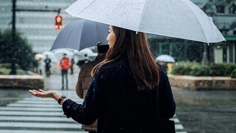 Mulher de costas segura guarda-chuva branco e estende a mão para sentir a neve que cai. Pessoas com guarda-chuvas ao fundo atravessam faixa de pedestres em área urbana.