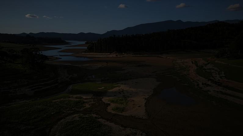 Represa apresenta grande extensão de solo exposto devido ao baixo nível de água, com pequenas poças remanescentes. Vegetação verde cerca a área, com colinas e montanhas ao fundo sob céu parcialmente nublado.