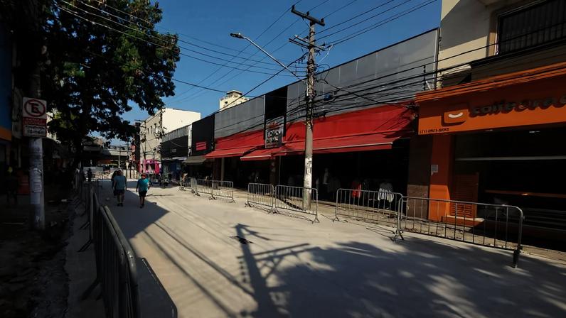 Rua comercial com lojas de fachadas coloridas à direita e à esquerda. Calçada à esquerda bloqueada por tapumes metálicos, com piso novo sendo instalado. Céu claro e árvores ao fundo.