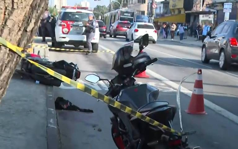 Motocicleta caída na calçada de uma rua movimentada, cercada por fita amarela de isolamento e cones de trânsito. Veículos e pedestres ao fundo, com viatura policial próxima ao local.