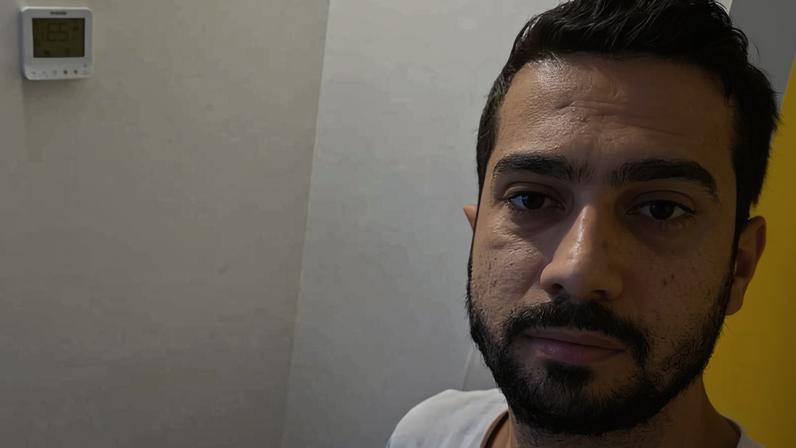 Homem de camiseta branca está ao lado de três malas grandes empilhadas em um canto de parede branca. Um termostato está fixado na parede acima das malas.