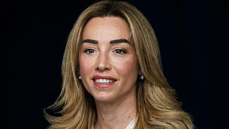 Silvia Salis sentada em uma mesa elegante, vestindo terno escuro com camisa azul clara, em ambiente interno sofisticado com luminária ao fundo.