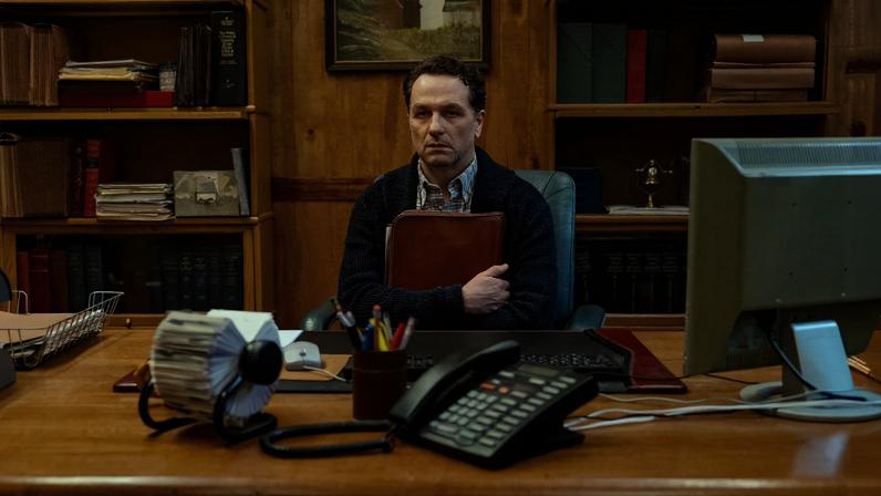 Homem sentado atrás de mesa de madeira em escritório com estantes cheias de livros ao fundo. Ele segura uma pasta marrom contra o peito e olha para frente. Na mesa há telefone, computador, porta-canetas e fichário giratório.