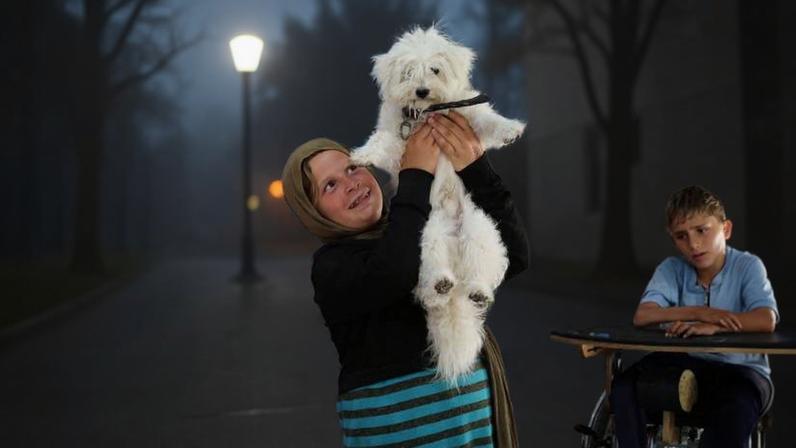 Menina com lenço na cabeça e vestido listrado segura um cachorro branco para cima em sala decorada com várias pinturas coloridas. Crianças sentadas observam ao redor.