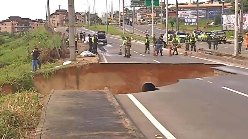 Grande deslizamento interrompe pista de rodovia urbana, com parte do asfalto desmoronada e buraco profundo. Equipe de emergência e policiais bloqueiam o trânsito no local, enquanto pedestres observam à margem da via.