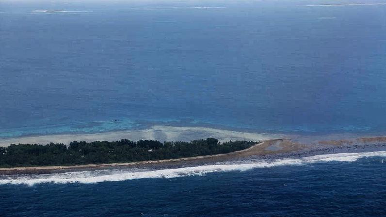 Vista aérea de uma ilha estreita com vegetação densa no centro, cercada por águas azuis do oceano. Ondas quebram na costa da ilha sob céu claro.