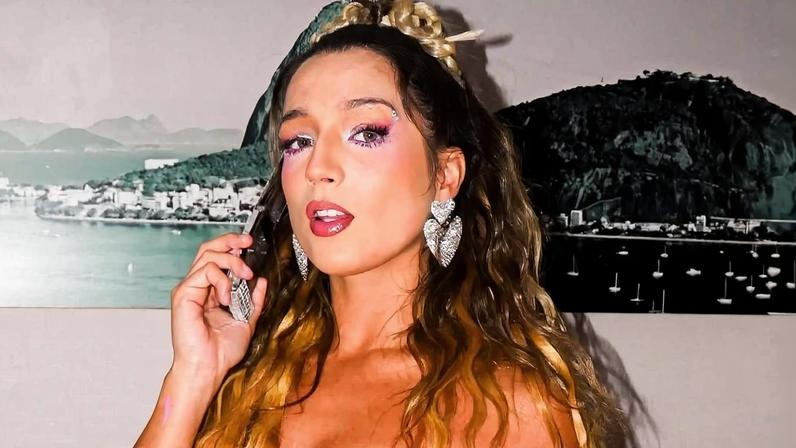 Mulher jovem com cabelo longo e ondulado, usando vestido rosa vibrante com detalhes brilhantes, posa em ambiente interno com parede clara e quadro preto e branco ao fundo.