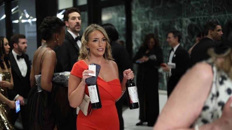 Mulher loira com vestido vermelho justo segura duas garrafas de vinho e um celular em evento formal com várias pessoas ao fundo vestidas com roupas de gala.