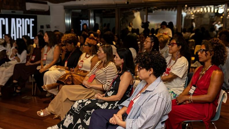 Grupo de mulheres sentadas em cadeiras alinhadas em ambiente interno, todas com crachás vermelhos pendurados no pescoço. Elas estão voltadas para a esquerda, aparentemente assistindo a uma apresentação. Ao fundo, há outras pessoas em pé e uma parede com a palavra 'CLARICE' iluminada.
