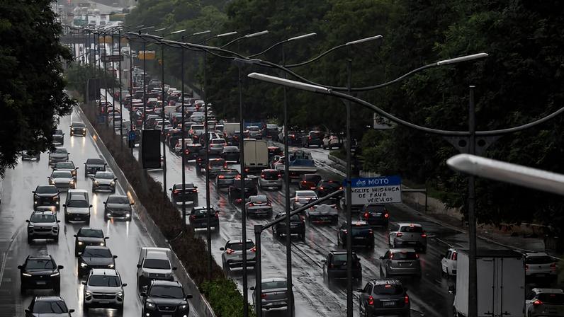 Congestionamento intenso na Marginal Pinheiros com veículos parados e lentos em várias faixas. Chuva deixa a pista molhada e reflexos visíveis no asfalto. Árvores e postes de iluminação acompanham a via.