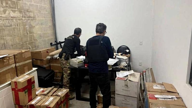 Dois homens de costas organizam documentos e caixas em sala pequena com paredes brancas e uma parede de blocos de concreto. A sala contém várias caixas de papelão empilhadas e uma mesa com papéis e equipamentos. Um aparelho de ar-condicionado está instalado na parede.