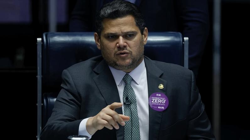Homem de terno escuro e gravata verde sentado em cadeira azul escura, gesticulando com a mão direita. Ele usa um botão roxo no paletó com texto sobre 'Princípio Zero'. Fundo desfocado com outra pessoa em pé.