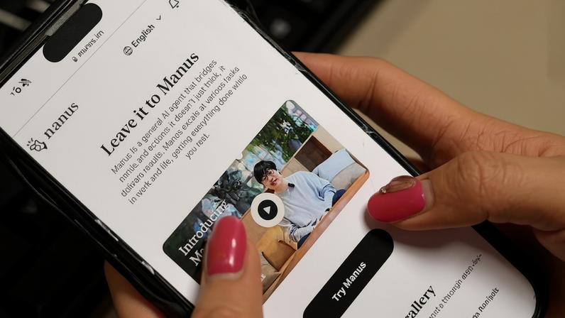 Mãos seguram smartphone exibindo página inicial do aplicativo Manus, com texto "Leave it to Manus" e botão "Try Manus". Tela mostra vídeo introdutório com homem sentado em sofá.