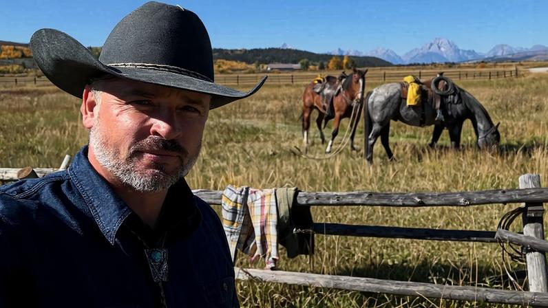Homem de meia-idade com chapéu de cowboy preto e camisa azul escura em primeiro plano, em campo aberto com vegetação seca. Ao fundo, três pessoas próximas a três cavalos equipados, com montanhas ao longe sob céu azul claro.