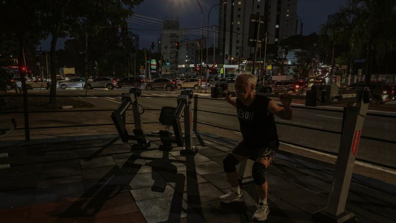 Homem idoso realiza agachamento em equipamento de academia ao ar livre próximo a avenida movimentada com carros e prédios ao fundo durante o anoitecer.