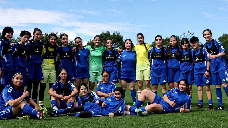 Vinte jogadoras de futebol feminino vestindo uniformes azuis e amarelos posam juntas no gramado de um campo ao ar livre sob céu azul com poucas nuvens.