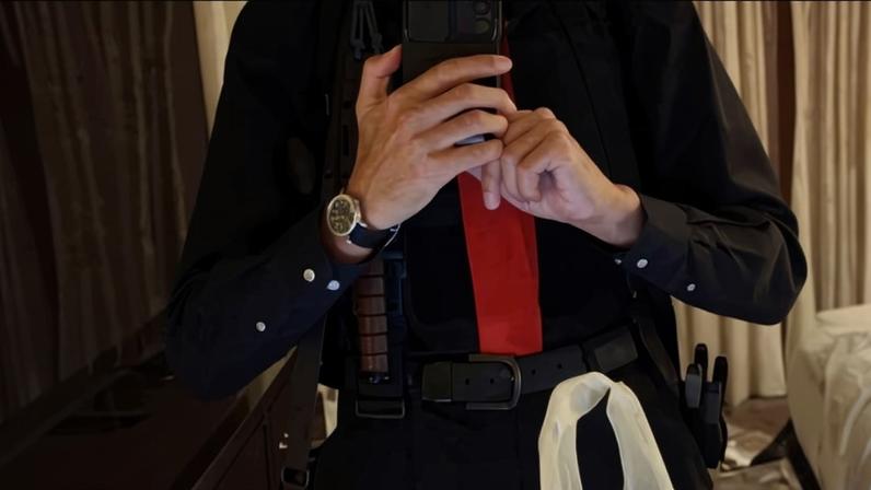 Homem com camisa preta e gravata vermelha tira selfie em espelho de quarto de hotel. Ele usa cinto com acessórios de segurança, incluindo algemas, rádio comunicador e caneta.