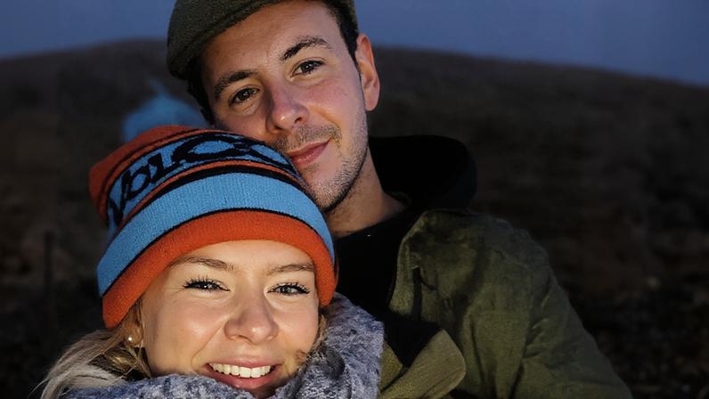 Homem e mulher posam juntos em ambiente externo com céu escurecendo ao fundo. Mulher usa gorro colorido azul e laranja e cachecol cinza, sorrindo. Homem atrás dela usa boné marrom e casaco verde, com expressão neutra.
