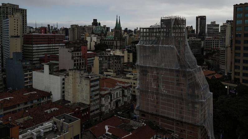 Vista aérea mostra prédios residenciais e comerciais em São Paulo sob céu nublado. Um edifício alto no centro está coberto por lona branca, indicando reforma. Ao fundo, destacam-se duas torres de igreja com cúpulas verdes.
