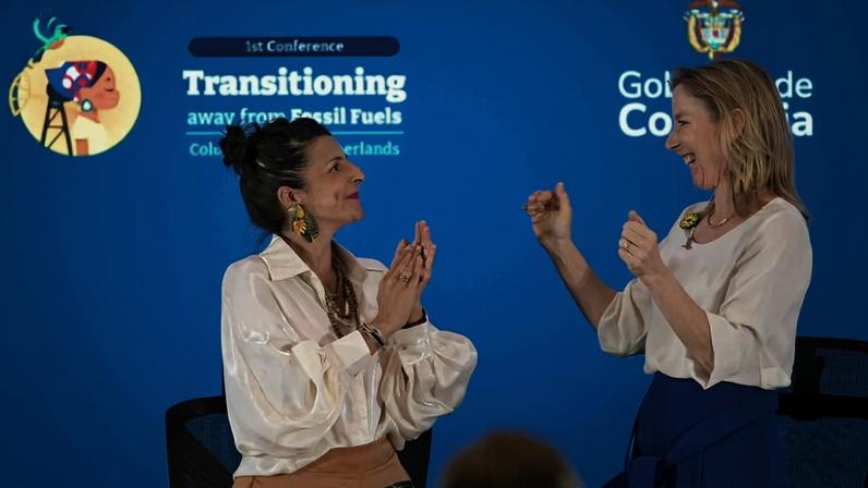 Duas mulheres de pé, uma com blusa branca e saia bege, outra com blusa branca e saia azul, trocam gestos de celebração em evento com fundo azul que exibe texto sobre transição de combustíveis fósseis e o governo da Colômbia.