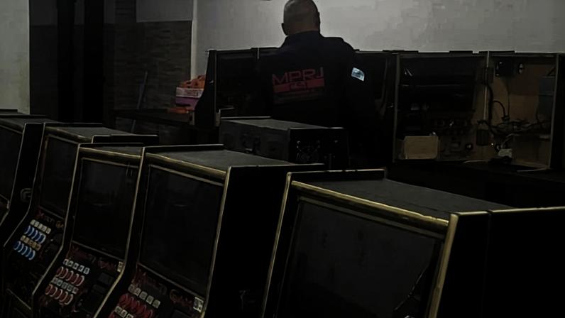 Homem de costas, vestindo camiseta preta com logo vermelho, operando máquinas caça-níqueis alinhadas em uma sala com paredes brancas e ventiladores de parede.