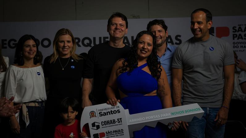 Dez adultos e uma criança posam em palco sob tenda branca. Mulher de vestido azul segura chave simbólica grande com logos de Santos, Conab e São Paulo. Pessoas ao redor sorriem e aplaudem. Fundo com banner do governo do estado de São Paulo.