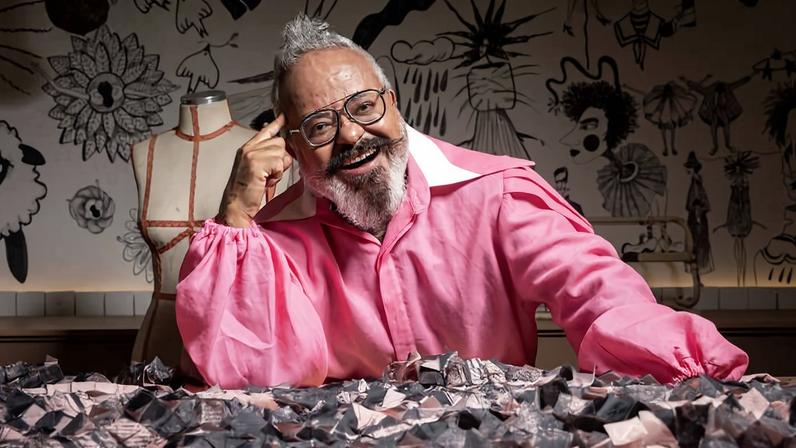 Homem de cabelos grisalhos e barba, usando óculos e camisa rosa, sorri enquanto apoia o braço sobre uma mesa coberta por tecido estampado. Ao fundo, manequim e parede decorada com desenhos artísticos em preto e branco.