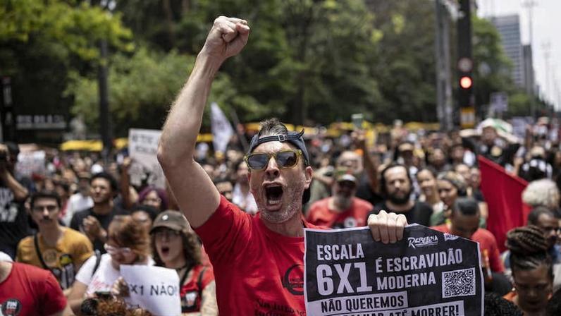Homem de camiseta vermelha e óculos escuros ergue o punho esquerdo e grita segurando cartaz contra escala 6x1, em meio a multidão de manifestantes na Avenida Paulista, com árvores e prédios ao fundo.