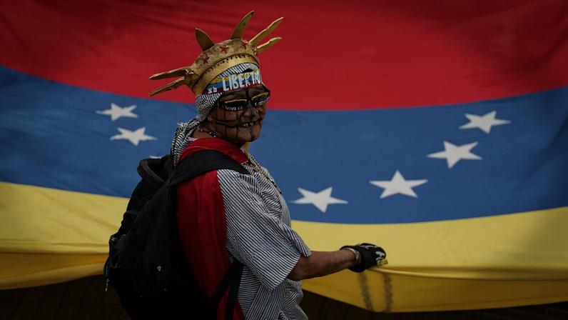Pessoa vestindo camiseta listrada e mochila preta segura a bandeira da Venezuela, que ocupa o fundo da imagem. Usa adereço dourado na cabeça com a palavra 'Libertad' e óculos escuros.