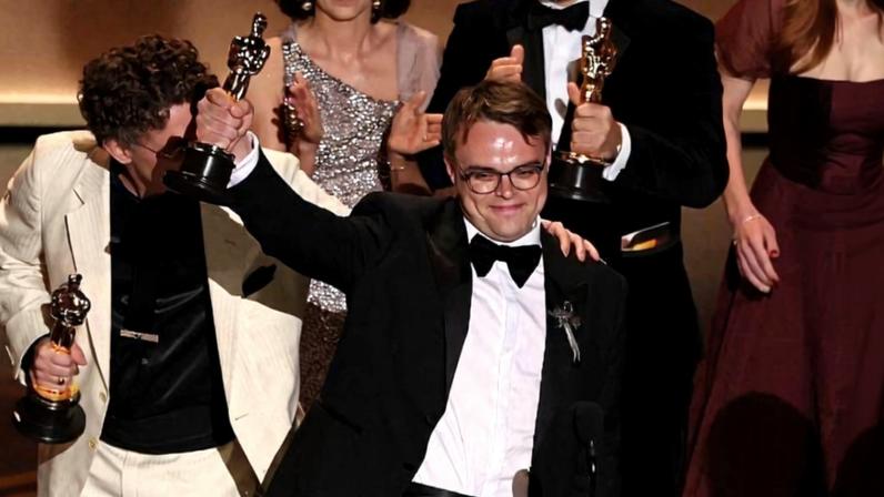 Seis pessoas vestidas formalmente posam no palco segurando estatuetas do Oscar. Um homem de terno preto levanta o braço com a estatueta, enquanto os demais sorriem e seguram seus prêmios.