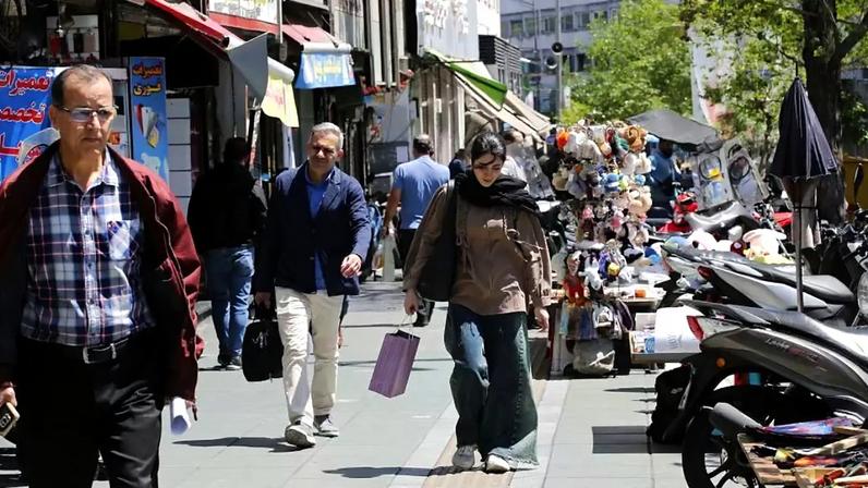 Pessoas caminhando em uma rua comercial movimentada em Teerã, com uma jovem carregando uma sacola de compras no centro da imagem.